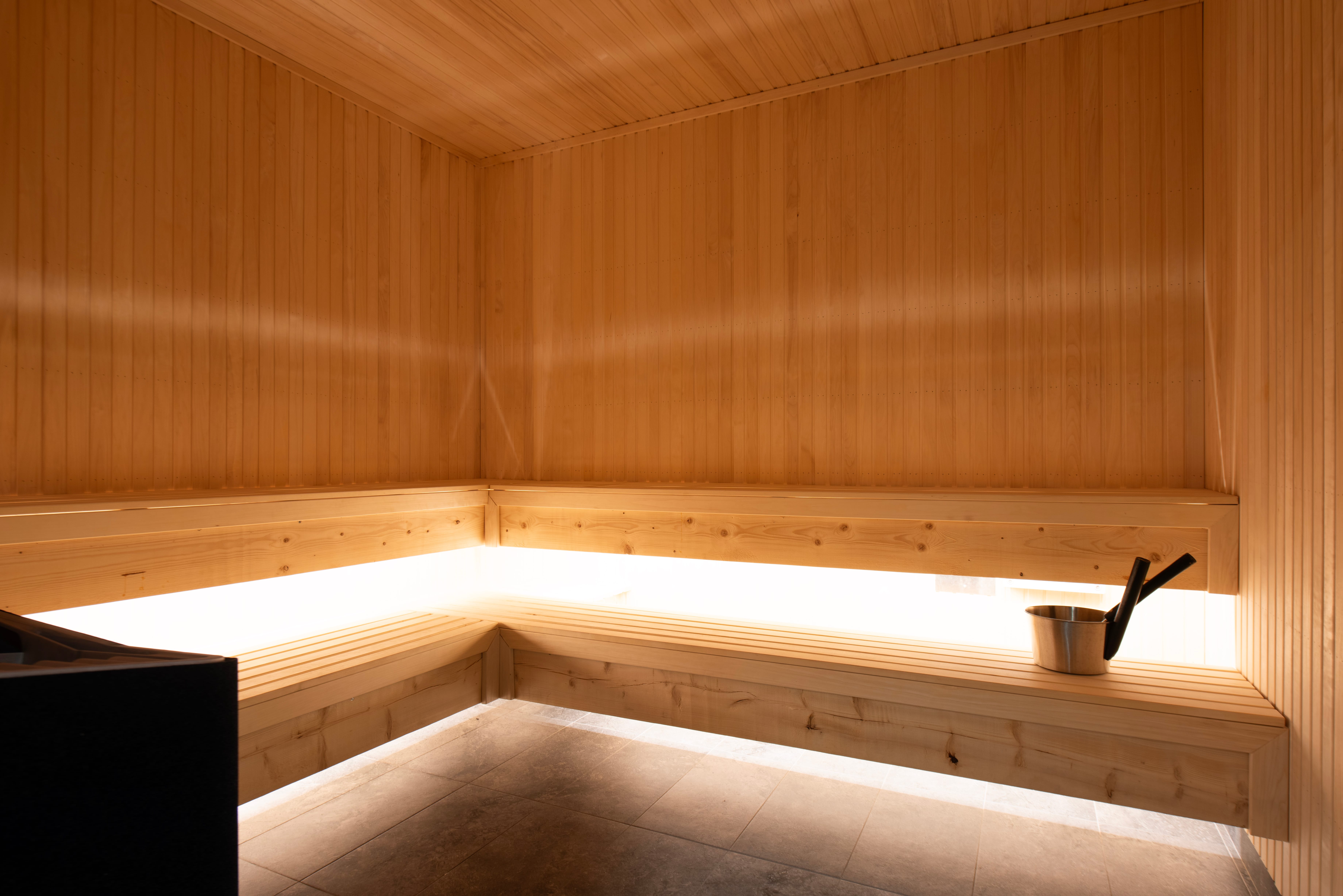 Bright wooden sauna with corner bench, stone wall floor and sauna bucket with ladle on the bench.