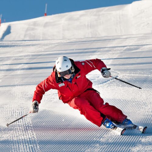 To skiløbere i røde dragter drejer hurtigt ned ad en præpareret piste, hvor sneen sprøjter.