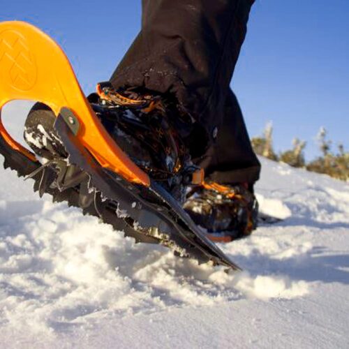 Close-up of orange snowshoes with metal spikes treading in sunlit snow during winter walk.