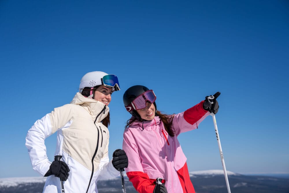 Två skidåkare med hjälm och goggles står i fjällandskap, en pekar framåt mot utsikt.