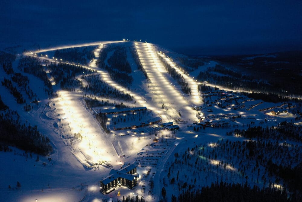 Oplyst skisportssted om natten med flere præparerede pister, lifte og bygninger i snedækket landskab.