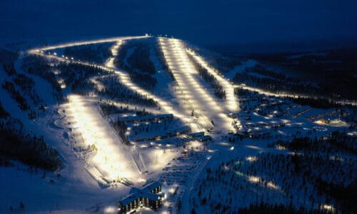 Upplyst skidort nattetid med flera preparerade backar, liftar och byggnader i snötäckt landskap.