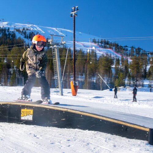 Person snowboarding on rail in sunny ski area with snow-covered slopes and blue sky