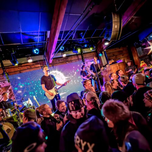 Livebands optræder på scenen med guitar, bas og trommer foran et stort publikum i et trælokale.