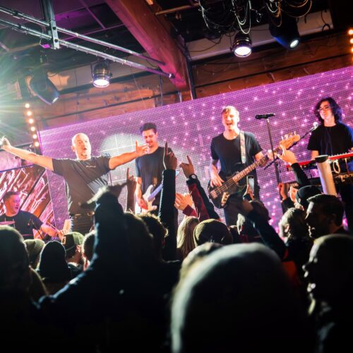 Live bands play on stage with guitars, bass and keyboards in front of an audience in a wood-panelled room with bright lighting.