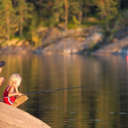 To børn sidder på en sten og fisker i en rolig sø med skov og klipper i baggrunden.