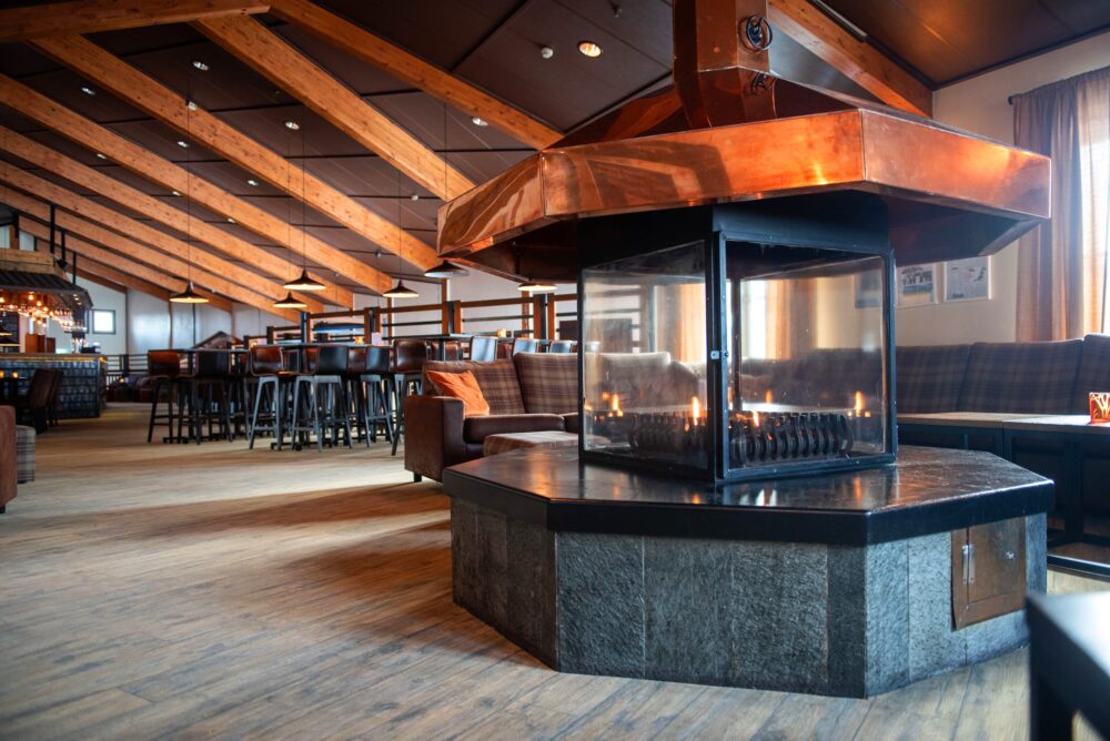 Stor lounge med centralt placerad öppen spis i koppar, omgiven av soffor och höga stolar.