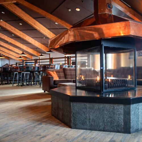 Stor opholdsstue med centralt placeret kobberpejs, omgivet af sofaer og høje stole.