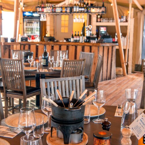 Rustikt restaurantinteriør med træbord, fonduegryde, vinglas og flasker, bar i baggrunden med hylder.