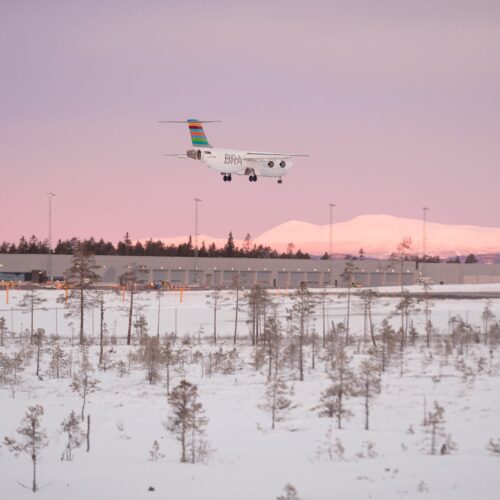 BRA-flygplan närmar sig Scandinavian Mountains Airport över snötäckt landskap vid rosa solnedgång.