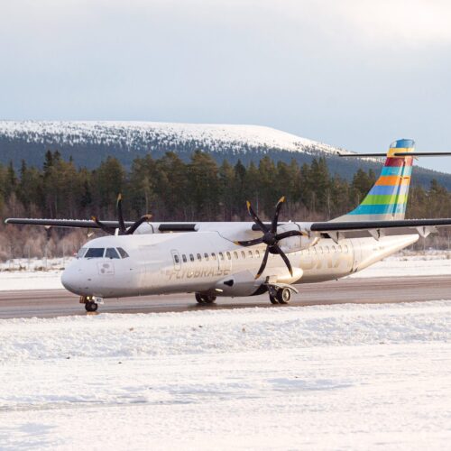 BRA propeller aircraft standing on snow-covered runway with forest and mountains in the background.
