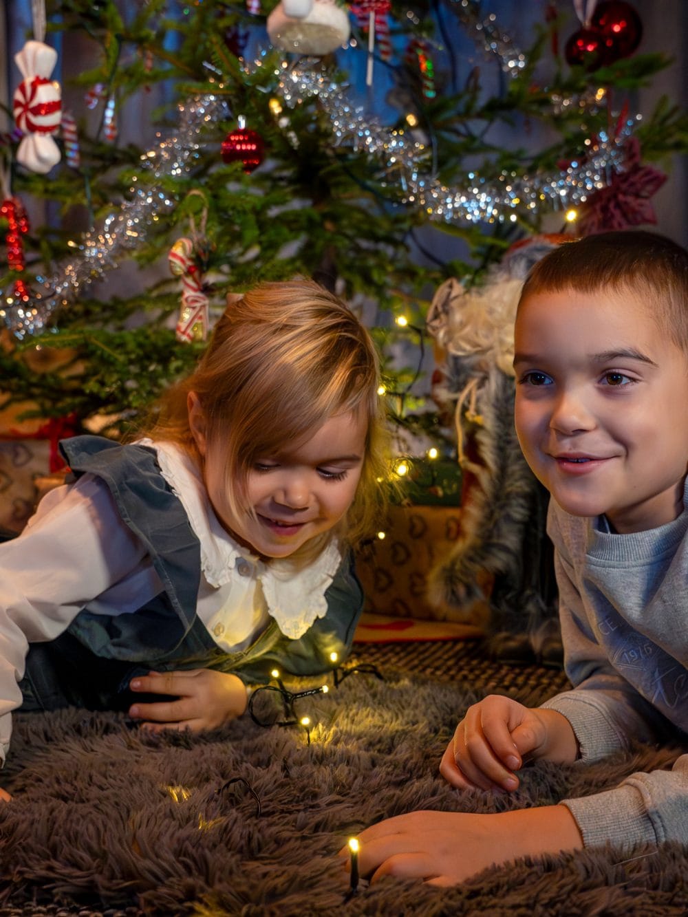 Två barn ligger på matta framför julgran med glitter och ljusslinga i stugan