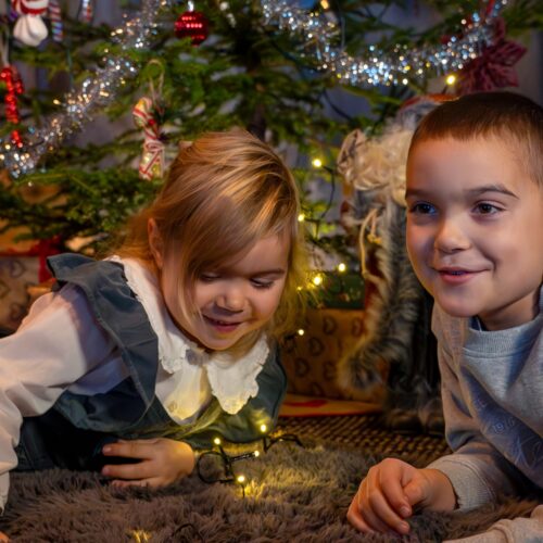 Två barn ligger på matta framför julgran med glitter och ljusslinga i stugan