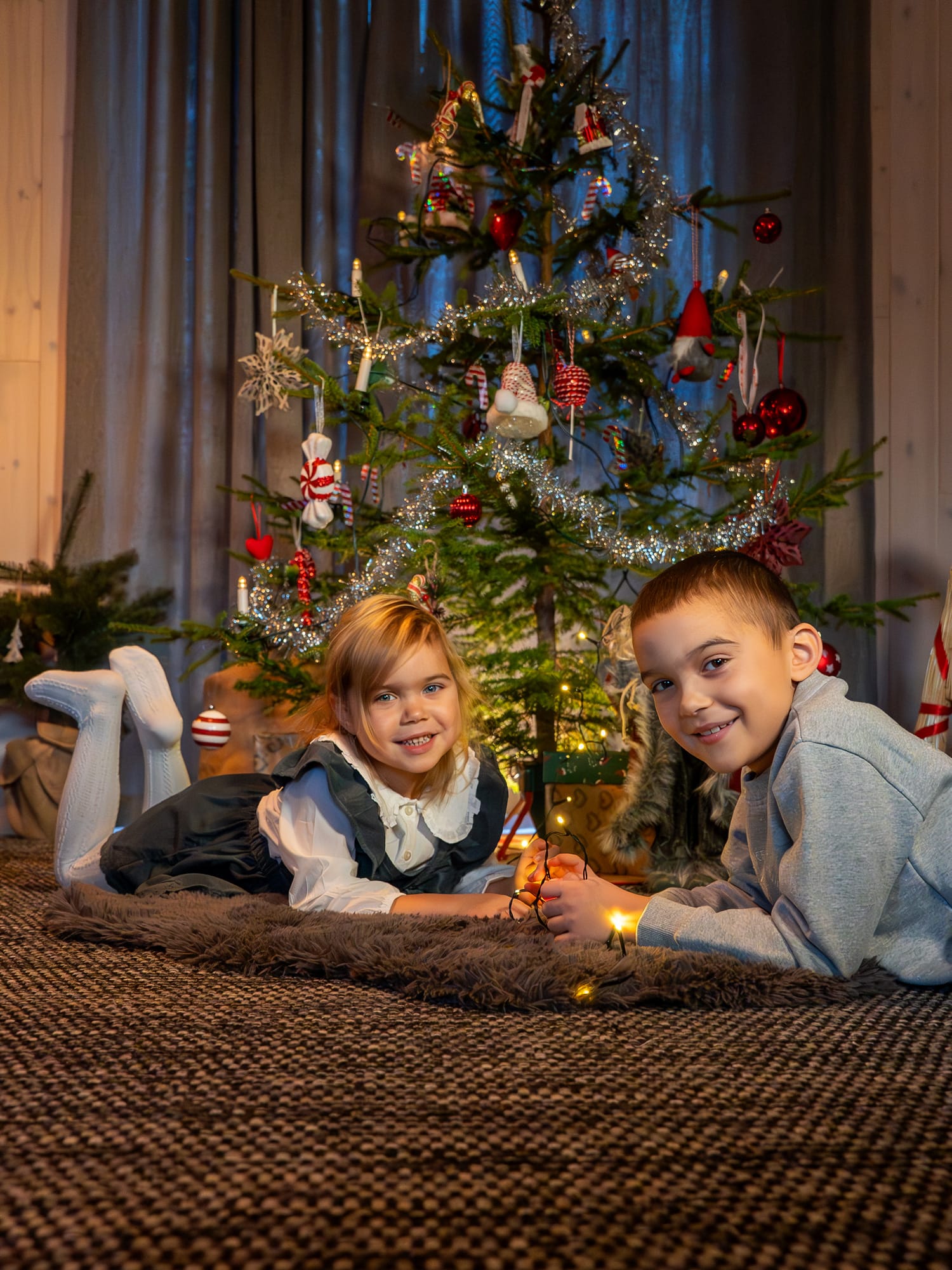 Två barn ligger på matta framför julgran med glitter, kulor och ljusslinga i stugan