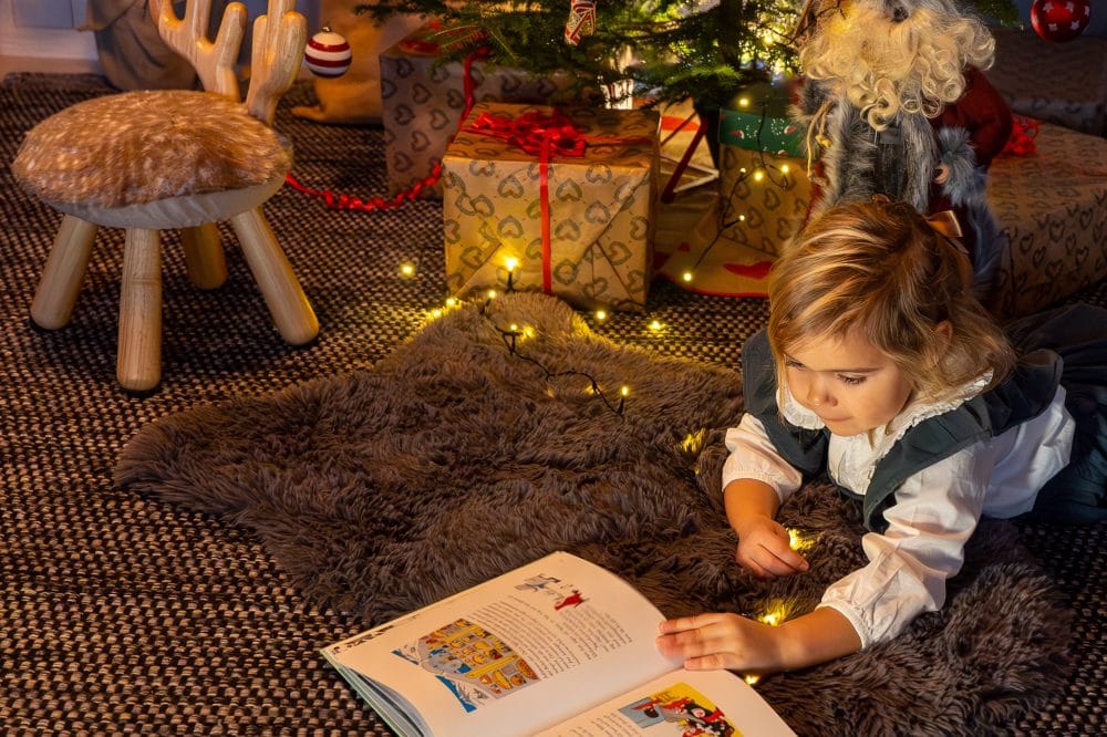 Children lying on a mat reading a book in front of a Christmas tree with tinsel and wrapped presents