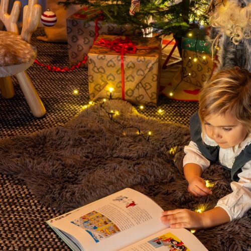 Barn ligger på matta och läser bok framför julgran med glitter och inslagna paket