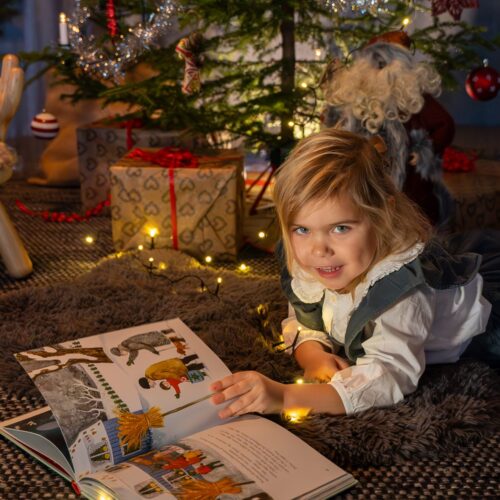 Barn ligger på matta och läser illustrerad bok framför julgran med paket, glitter och halmbock