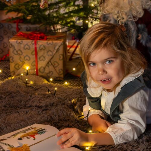 Barn ligger på matta och läser bok framför julgran med glitter och inslagna paket