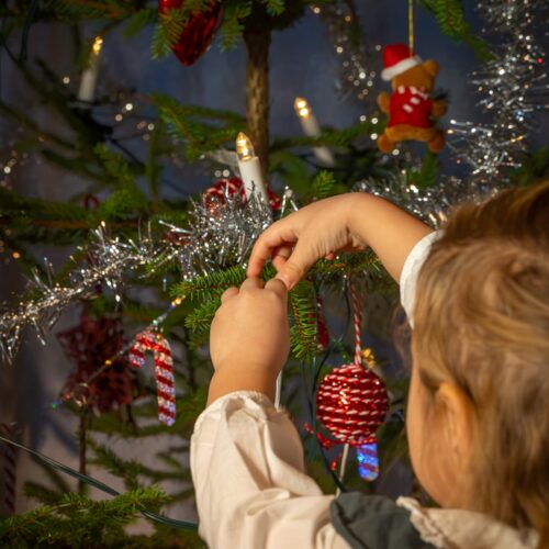 Barn hänger glittergirlang på julgran dekorerad med röda kulor, polkagrisar och elektriska ljus, med inslagna paket under granen