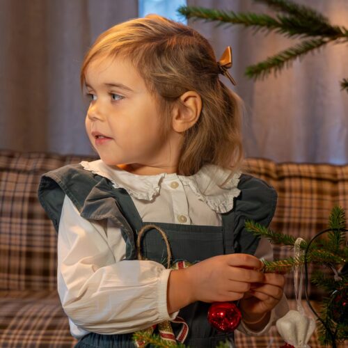 Barn klär julgran med röda kulor och hjärtformad dekoration, framför soffa med rutigt tyg