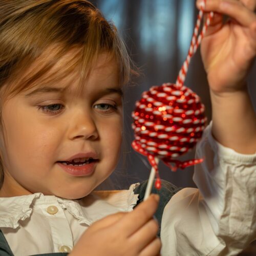 Barn håller upp rödvit julgranskula med snöre framför julgran i stämningsfull miljö