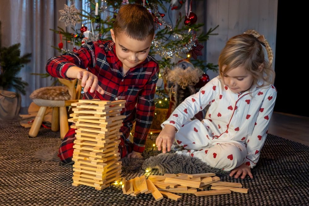 Två barn i pyjamas bygger högt torn av träklossar framför julgran med glitter och halmbock