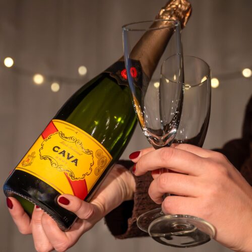 Cava bottle with yellow label held together with two champagne glasses against a background of string lights
