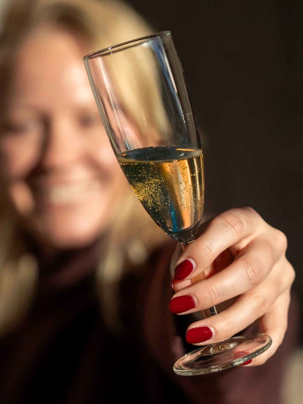 Hand med rödmålade naglar håller upp glas med mousserande vin och bubblor mot mörk bakgrund