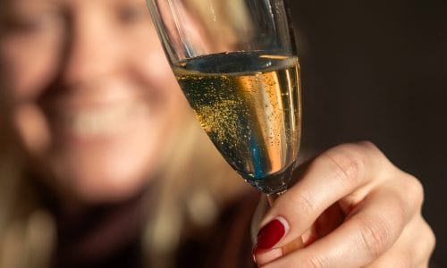 Hånd med rødmalede negle holder glas med mousserende vin og bobler op mod mørk baggrund