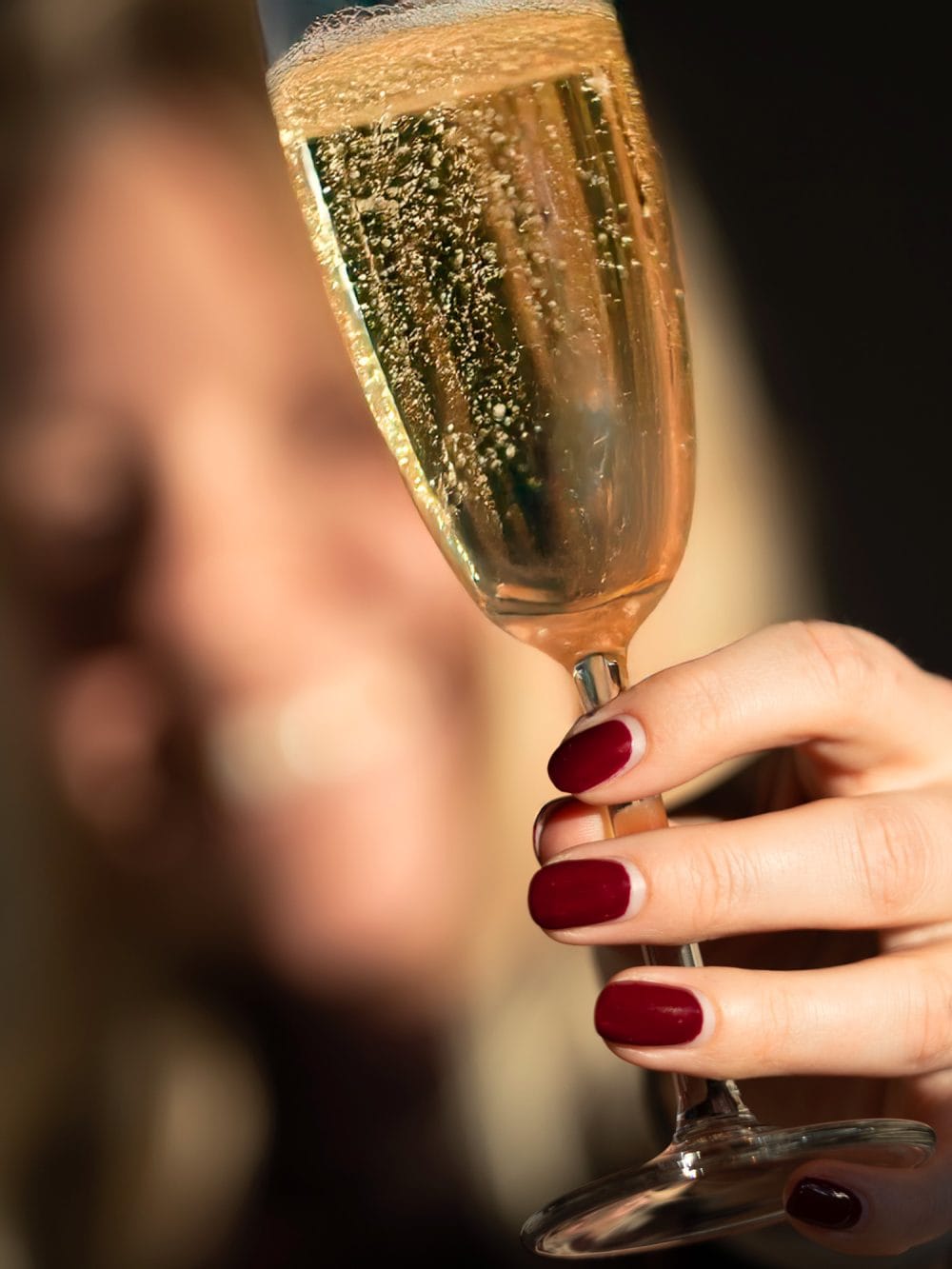 Hand med rödmålade naglar håller upp glas med mousserande vin och bubblor mot mörk bakgrund