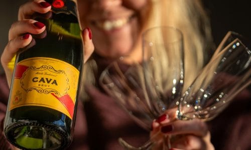 Cava bottle with yellow label held next to three empty champagne glasses against dark background