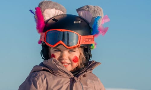 Child in pink jacket with black helmet decorated with colourful feathers and grey plush ears against blue sky