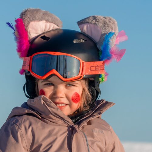 Barn i rosa jacka med svart hjälm dekorerad med färgglada fjädrar och grå plyschöron mot blå himmel