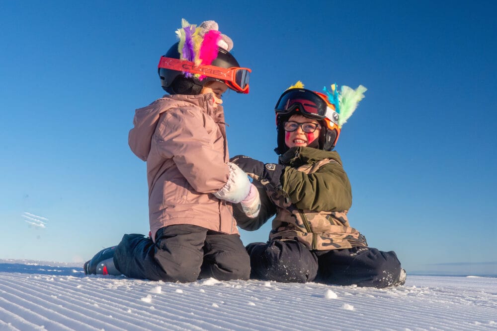 To børn i skidragter og hjelme med farverige påskefjer sidder på et snedækket bjerg under en blå himmel.
