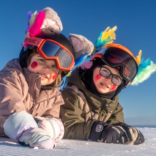 Two children in ski suits and helmets with colourful Easter feathers lie on a snowy mountain under a blue sky