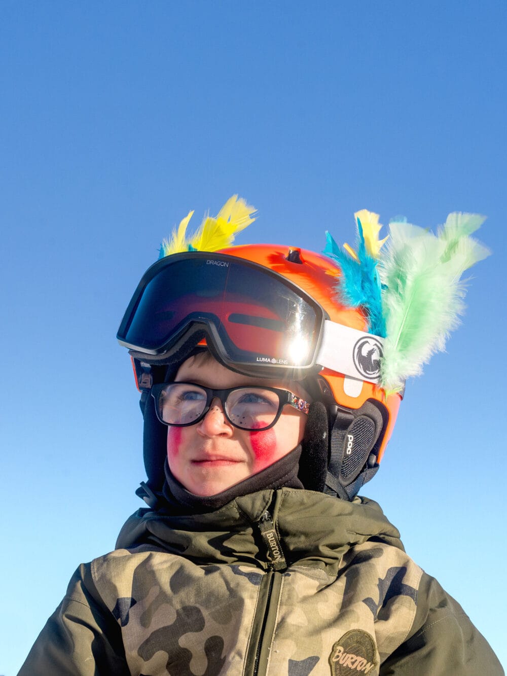 Barn i mørk jakke med orange hjelm dekoreret med farverige fjer mod blå himmel