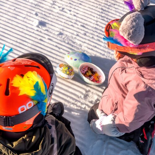 Två barn i skidkläder sitter på snö bredvid öppna påskägg fyllda med godis