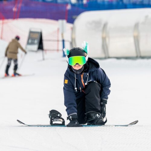 Børn snapper snowboardbindinger på en snedækket skråning, hjelme med farverige påskefjer og skiløbere i baggrunden.