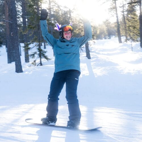 En person på snowboard med blå jakke og farverig hjelmdekoration kører på en solbeskinnet skovbakke.