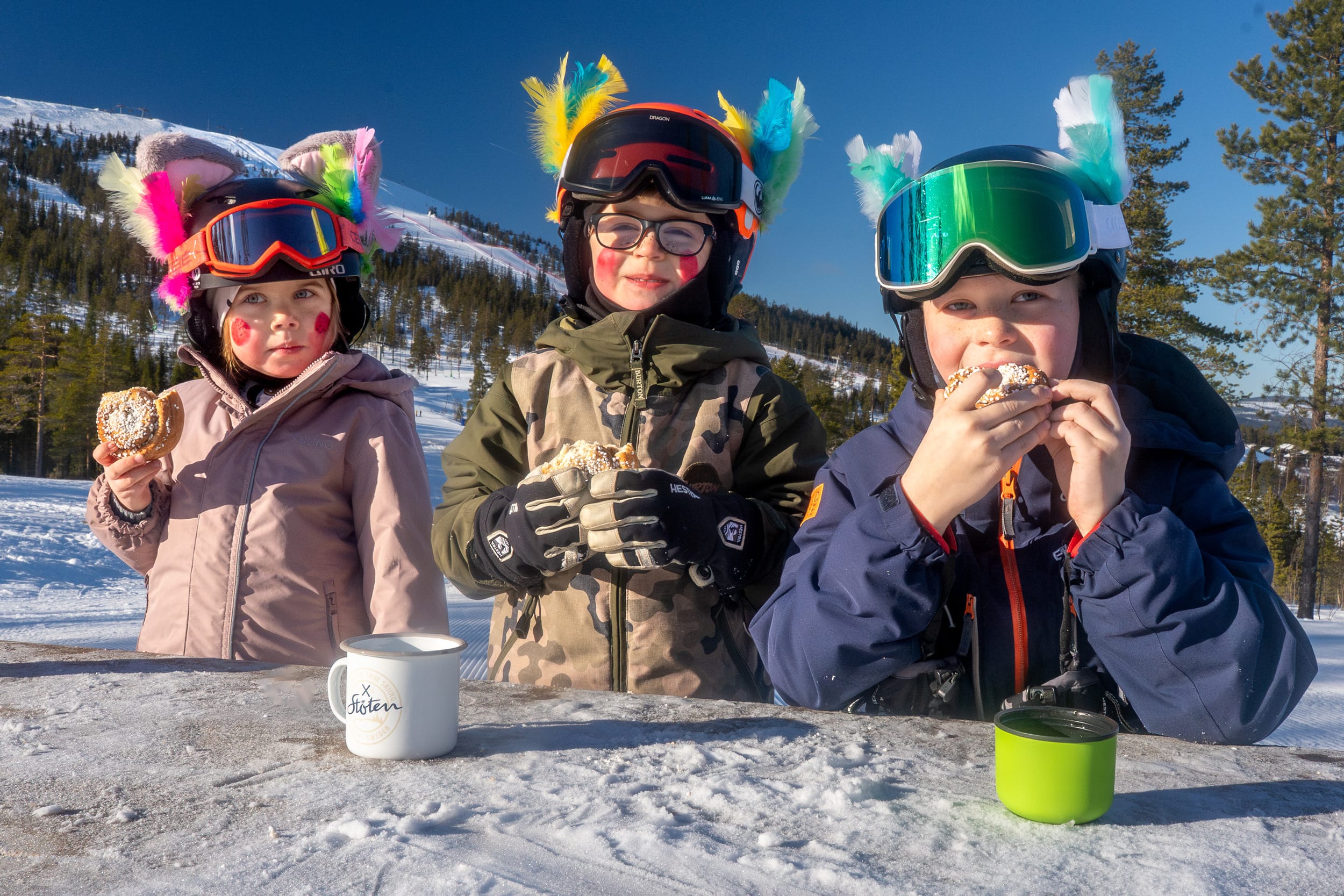 Tre barn i skidkläder och hjälmar med färgglada dekorationer fikar vid träbord på fjället