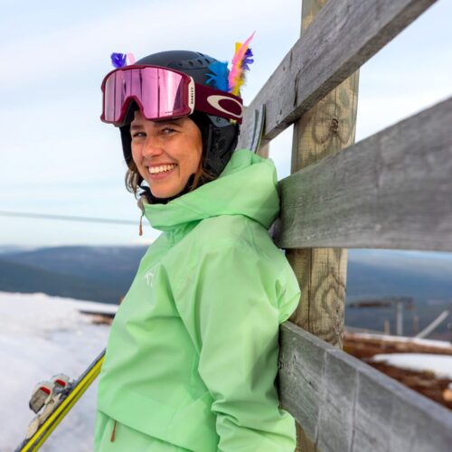 Person i grøn skijakke med ski læner sig op ad træhegn, bjerge og lift i baggrunden