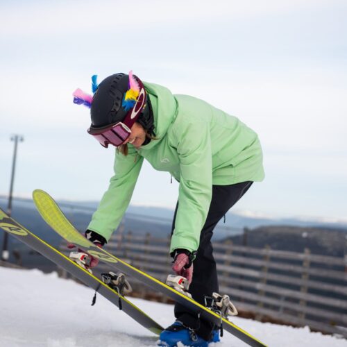 Person in green ski jacket standing on snowy slope holding two skis, helmet with colourful Easter feathers