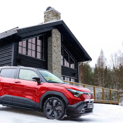 Red Suzuki Vitara parked in the snow in front of a modern mountain cabin with black wooden facade and stone-walled chimney.