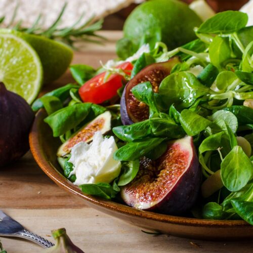 Grøn salat med friske figner, tomater, lime og krydderurter i rustik skål på træbord.