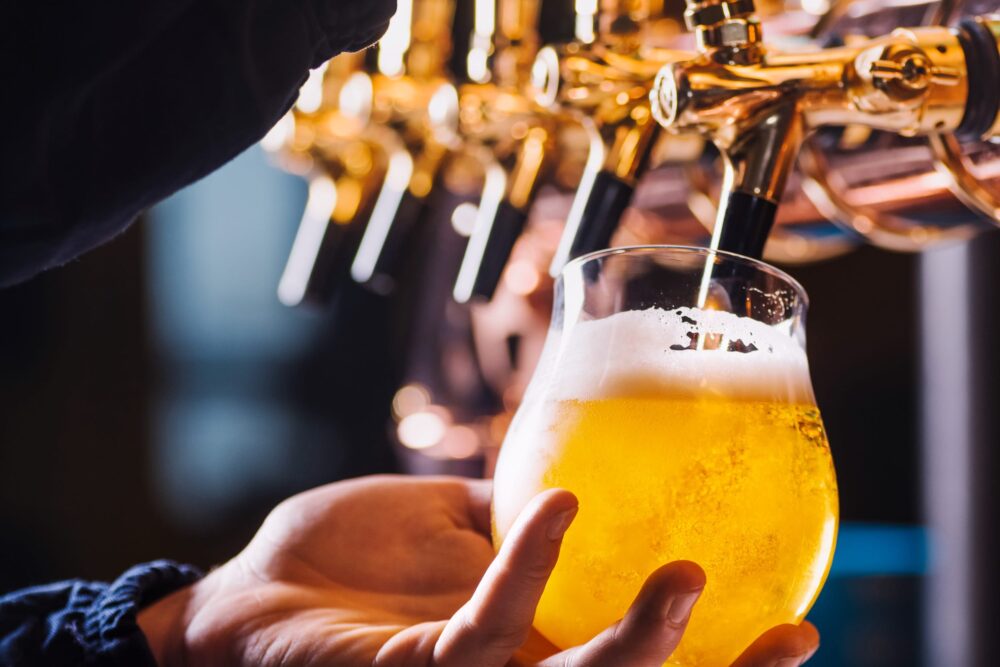 Hand håller glas fyllt med gyllene öl under tappkran i bar med mässingsdetaljer.