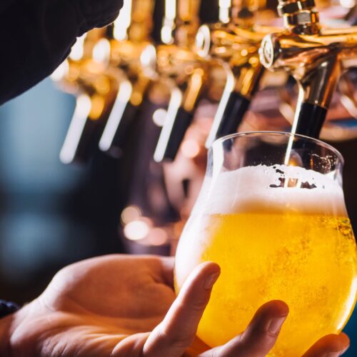 Hand holds glass filled with golden beer under tap in bar with brass details.