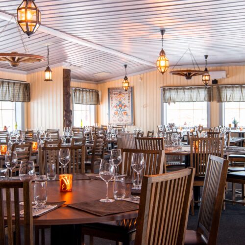 Stor spisestue med træborde, vinglas, tændte lanterner og rustikke lamper i loftet.