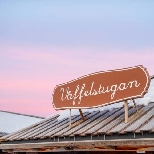 Skylt med texten “Våffelstugan” på snötäckt tak mot rosa och blå vinterhimmel.
