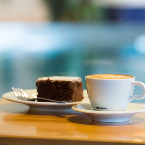 Kaffekop med cappuccino og chokoladekage på bakke ved træbord nær poolområdet.
