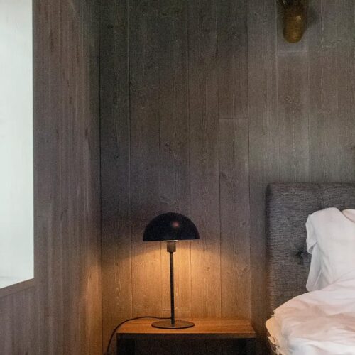 Mörk träpanelvägg med säng i grått tyg, vita sängkläder, svart bordslampa på träbord och grön växt i fönsterkarm.